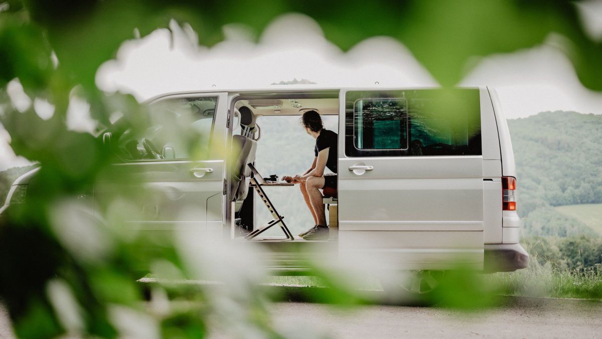 Ein Van parkt neben einer hochgelegenen Landstraße, durch die offenen Schiebetüren sieht man drinnen einen jungen Mann beim Laptop sitzen. Im Hintergrund ein schöner Ausblick auf die umliegende Landschaft.