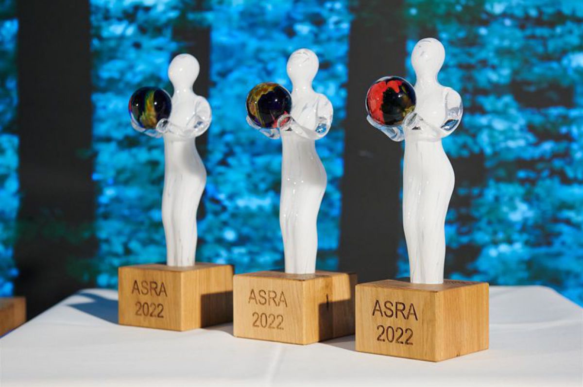 Der ASRA Preis: Eine weiße Glasfigur auf hölzernem Sockel hält eine bunte Glasmurmel als Symbol für die Erde in Händen. Auf dem Sockel die Schrift 
