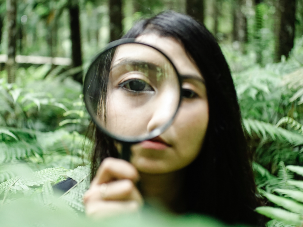 Ein Mädchen steht schulterhoch inmitten von Farnen in einem Wald und hält sich eine Lupe vors Gesicht, sodass man ihr rechtes Auge vergrößert im Zentrum des Bildes sieht.