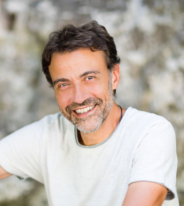 Peter Schramek: Ein Mann mit kurzem Haarschnitt und ergrauendem Bart lächelt in die Kamera. Er trägt ein weißes T-Shirt und sitzt unter einem Baum, im Hintergrund eine Felswand.