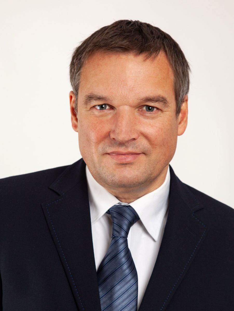 Unternehmensberater Albert Huber hat kurze braune Haare und ein glattrasiertes Gesicht. Er trägt einen schwarzen Anzug, weißes Hemd und eine dunkelblaue Krawatte. Der Bildhintergrund ist weiß.