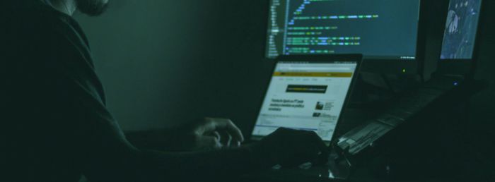 In einem dunklen Raum, nur schwach vom grünlichen Licht der Bildschirme erhellt, sitzt ein Mann mit Bart und Brille vor einem Laptop und mehrern Monitoren in verschiedenen Größen. Auf dem Hauptbildschirm sieht man lange hellgrüne Buchstaben- und Zahlenrei
