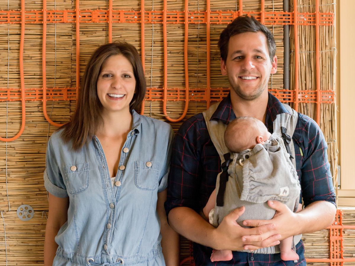 Victoria und Lukas mit Baby Emil vor einer mit Stroh gedämmten Wand mit orangen Heizschläuchen. Vater Lukas trägt seinen Sohn in einer Tragehilfe vor dem Oberkörper.