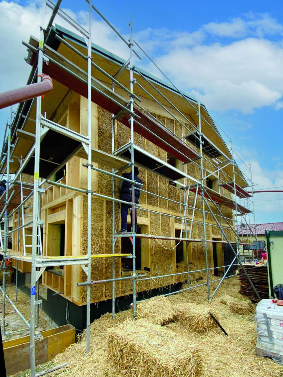 Das Strohgedämmte Haus mit Dach und Gerüsten vor den Außenwänden. Auf der Baustelle rund um das Haus liegen eckige Strohballen.