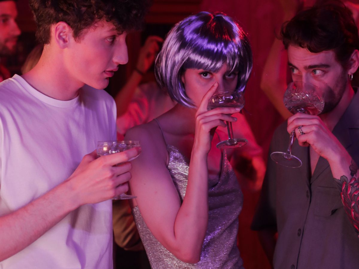 Drei Jugendliche im Fokus beim Feiern auf einer Wohnzimmerparty. Die junge Frau in der Mitte trägt ein enges Glitzerkleid und eine lila Pagenkopfperücke. Links und rechts von ihr jeweils ein junger Mann, alle drei trinken aus Sektgläsern. Das Licht im Rau