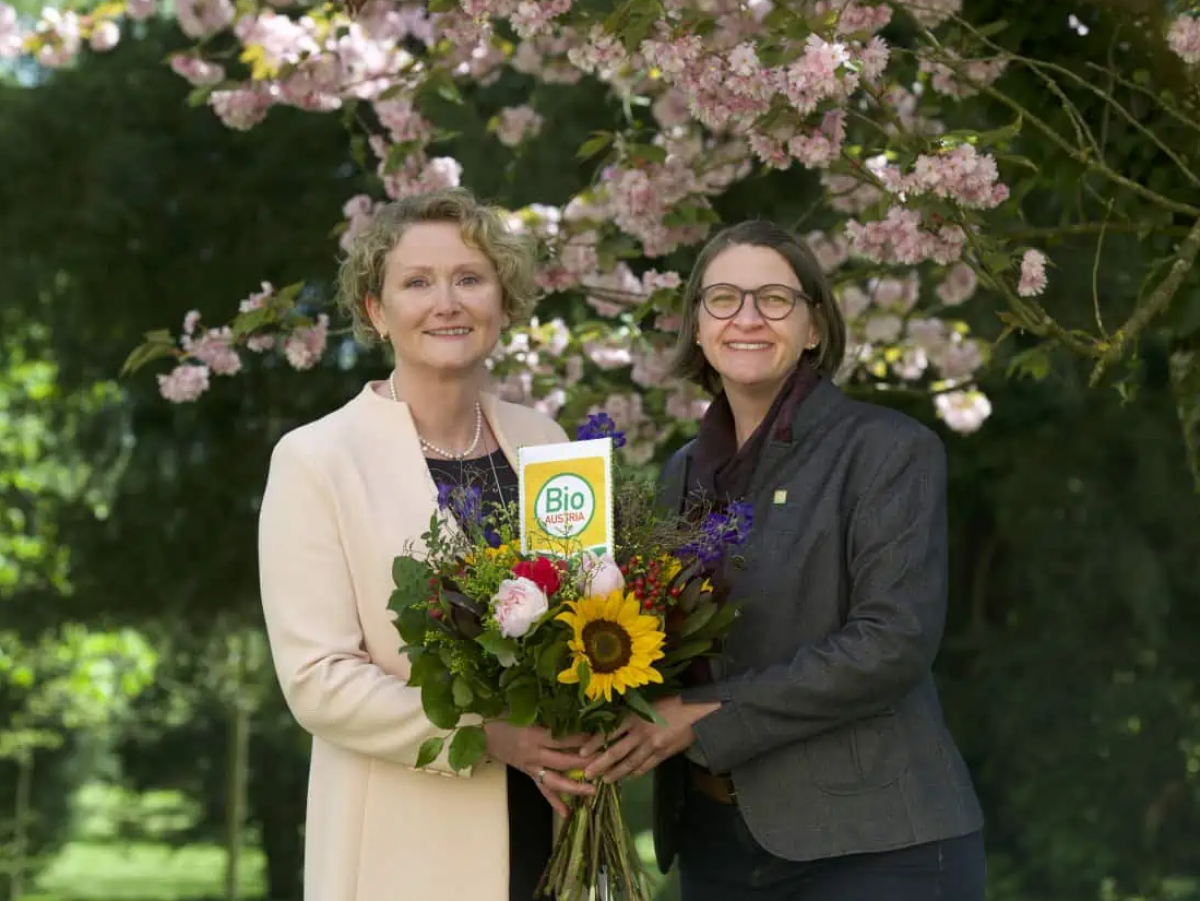 Altobfrau Gertraud Grabner und die neu gewählte Obfrau Barbara Riegler stehen vor einem blühenden Obstbaum, halten zusammen einen bunten Blumenstrauß und lächeln in die Kamera.