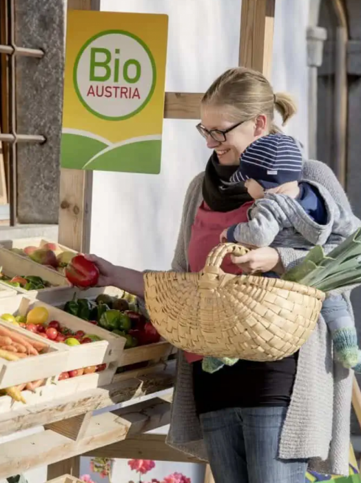 Eine Mutter mit Baby auf dem Arm und Einkaufskorb in derselben Hand nimmt gerade eine rote Paprila aus einer Holzsteige an einem Bio-Gemüsestand. Das Logo von BIO AUSTRIA hängt an der Holzwand.