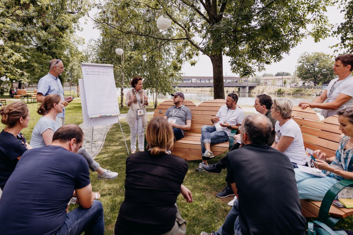 Menschen sitzen im Freien, im Kreis und diskutieren. Weiters ist ein Flipchart zu sehen, auf dem die Ergebnisse notiert werden.