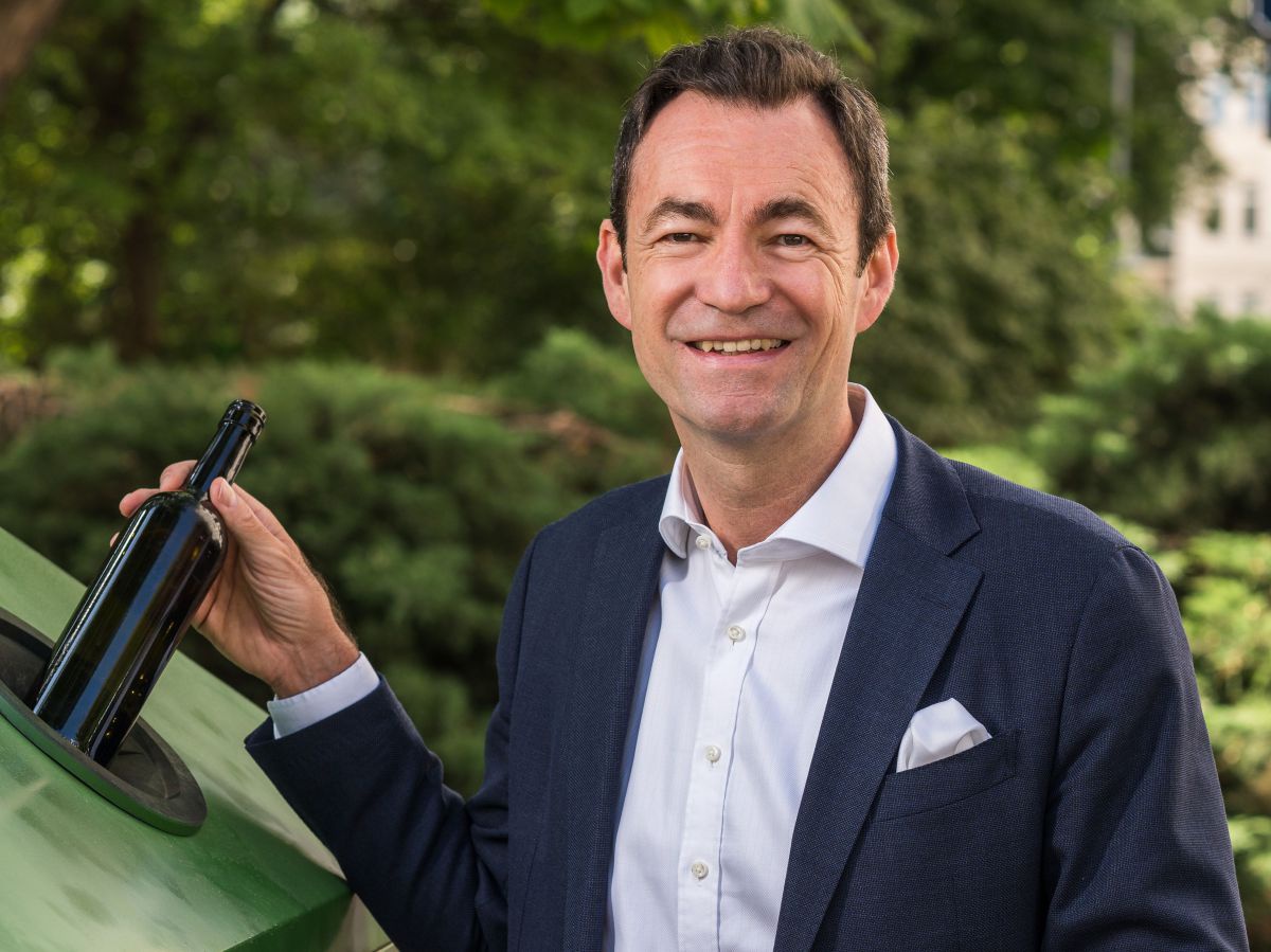 Glasrecycling ist Klimaschutz: Dr. Harald Hauke, Geschäftsführer der Austria Glas Recycling GmbH, steht in weißem Hemd und Business-Anzug an einem der neuen Sammelcontainer und wirft lächelnd eine dunkle Weinflasche in die Öffnung für Buntglas.