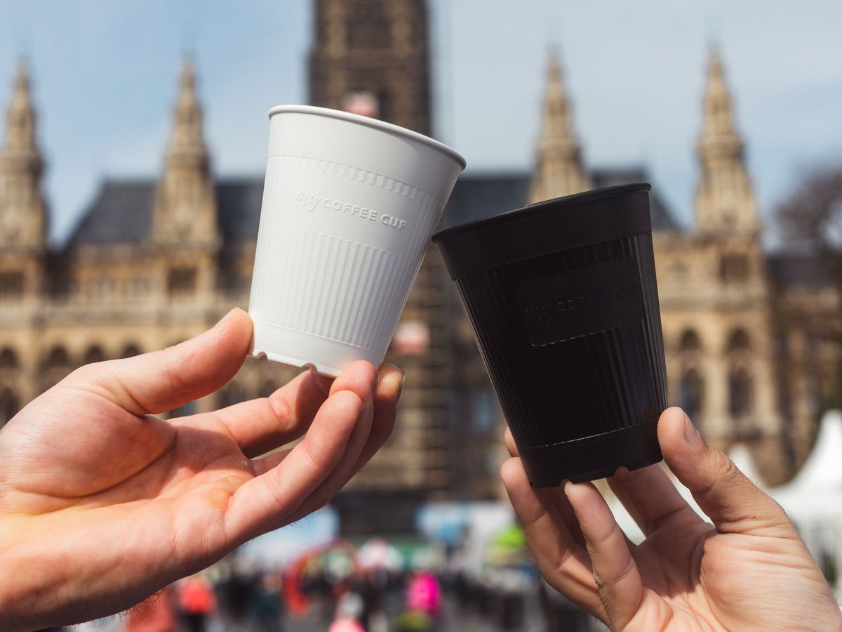 Zwei Hände halten Kaffebecher zusammen, als würden sie sich zuprosten. Im Hintergrund das Wiener Rathaus.