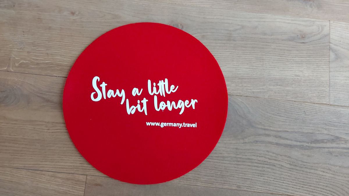 Ein rotes Sitzkissen liegt auf einer Holzbank. Auf dem Kissen steht: Stay-a-little-bit-longer in weißer Schrift.