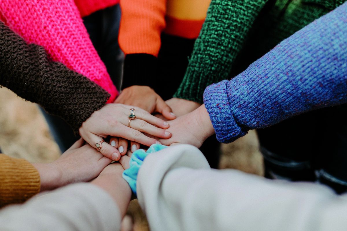 Acht junge Menschen stehen zusammen im Kreis und legen ihre Hände aufeinander. Die unterschiedlichen Farben der Ärmel ergeben ein buntes Bild.