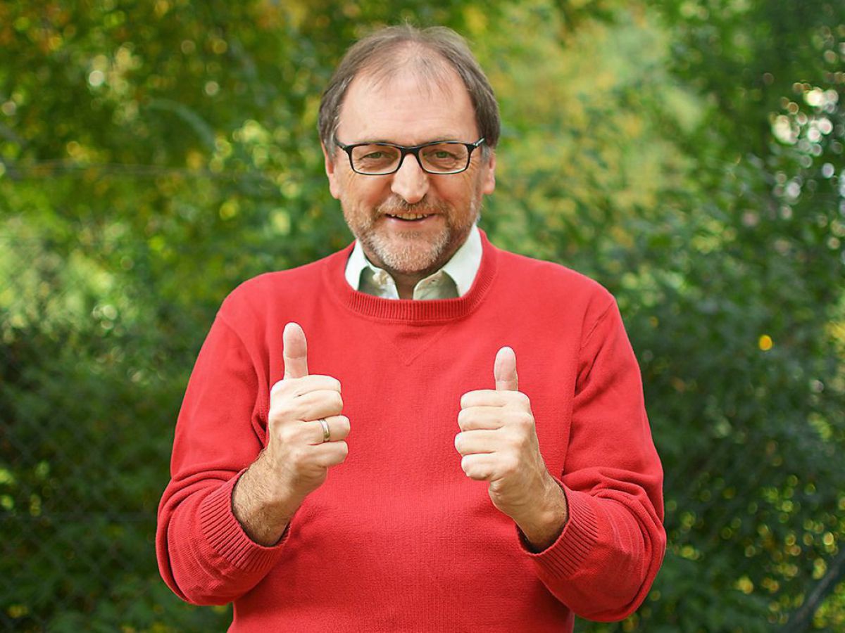 Reinhard Heiserer im roten Pullover vor blattgrünem Hintergrund zeigt lächelnd beide Daumen hoch.