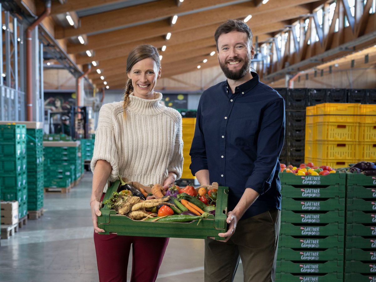 Cornelia und Andreas Diesenreiter stehen lächelnd in einer hellen Lagerhalle in der Kisten voller Obst und Gemüse gestapelt sind und halten gemeinsam eine grüne Kartonsteige voller Gemüse.