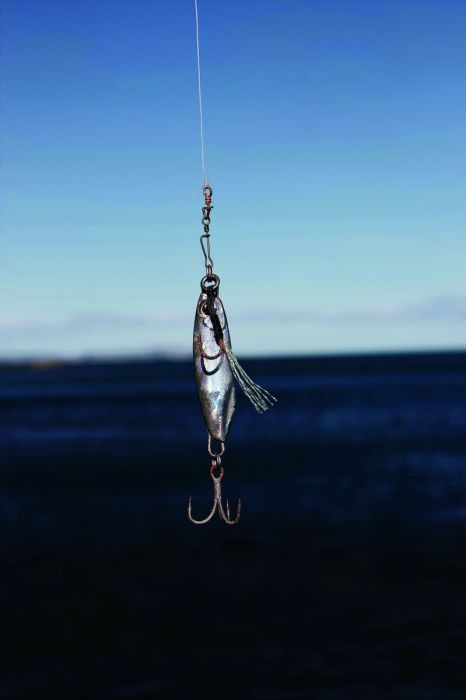 Ein Angelhaken mit Köder in Form eines kleinen silbernen Fisches hängt an einer Angelschnur. Im Hintergrund blauer Himmel über dunkelblauem Meer.