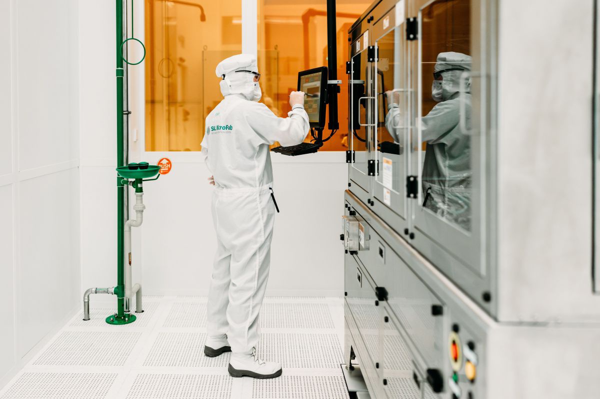 Ein*e Labortechniker*in im weißen Schutzanzug steht in einem grellweißen Laborraum vor Schränken mit Glastüren und hantiert mit einem Touchsreen.