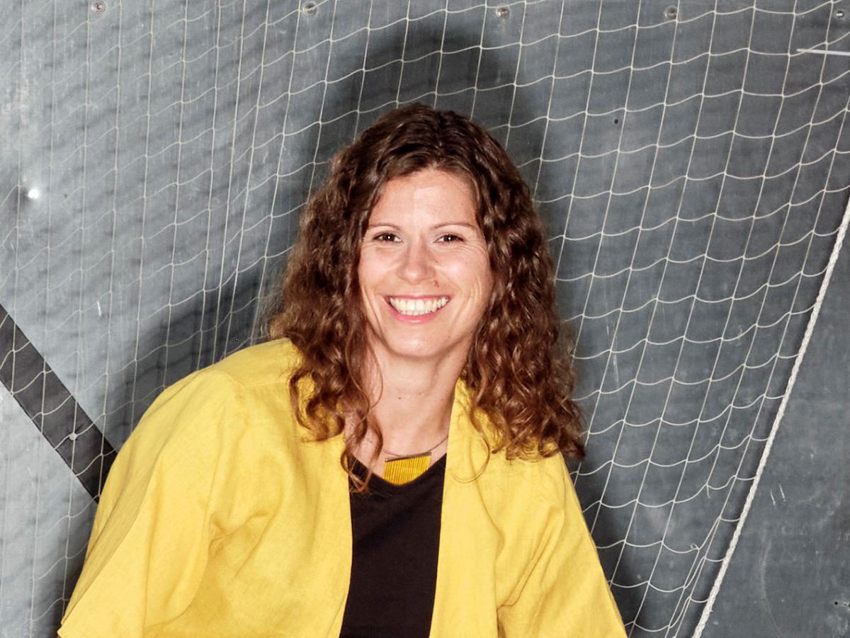 Jugendlich wirkende Frau mit dunkelblonden, schulterlangen Locken. Sie trägt eine knallgelbe, kurzärmelige Hemdjacke über schwarzem T-Shirt und strahlt in die Kamera.