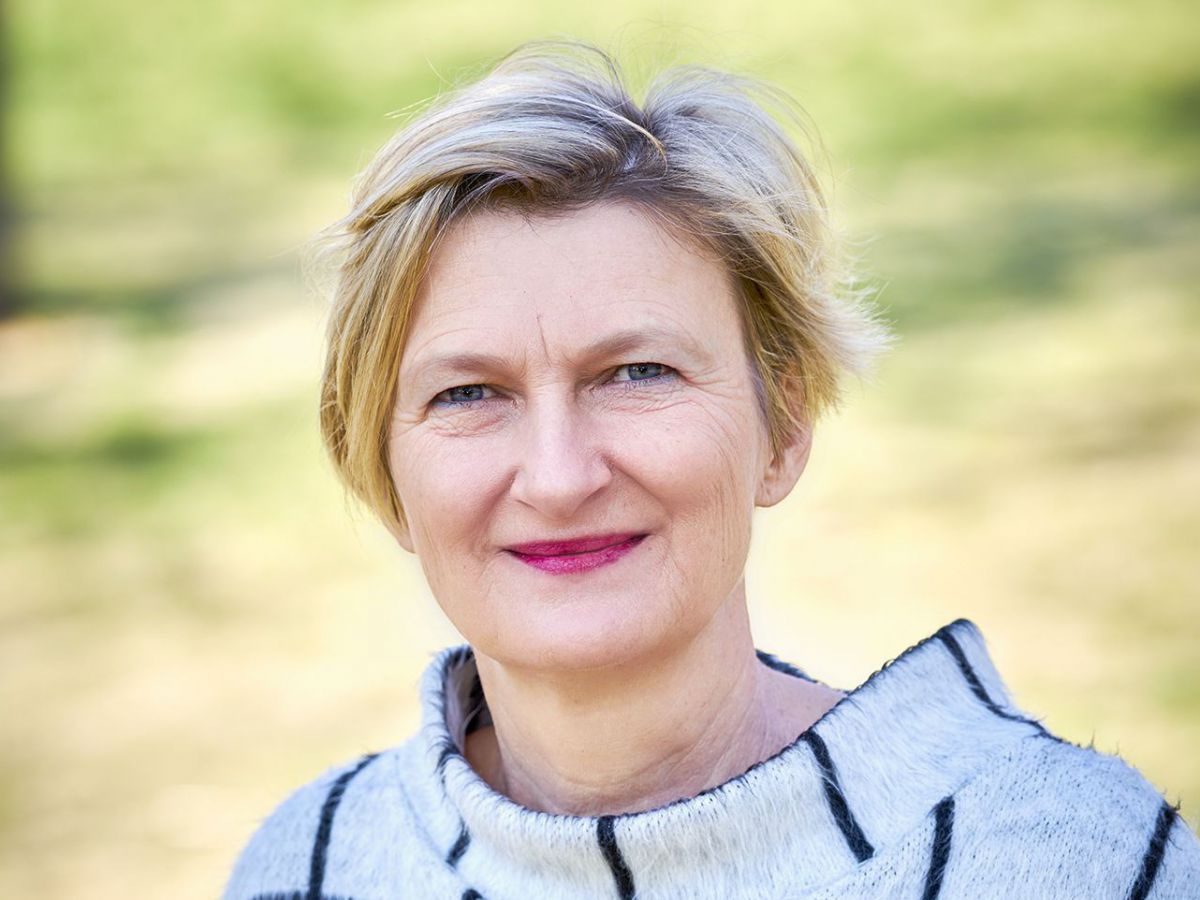 Frau mit blondem Pagenschnitt, blauen Augen und rotem Lippenstift, sie trägt ein naturweißes Wollkostüm mit schwarzem, großmaschigem Karomuster. Das Foto wurde im Freien aufgenommen, der Wind bläst leicht durch ihr Haar.