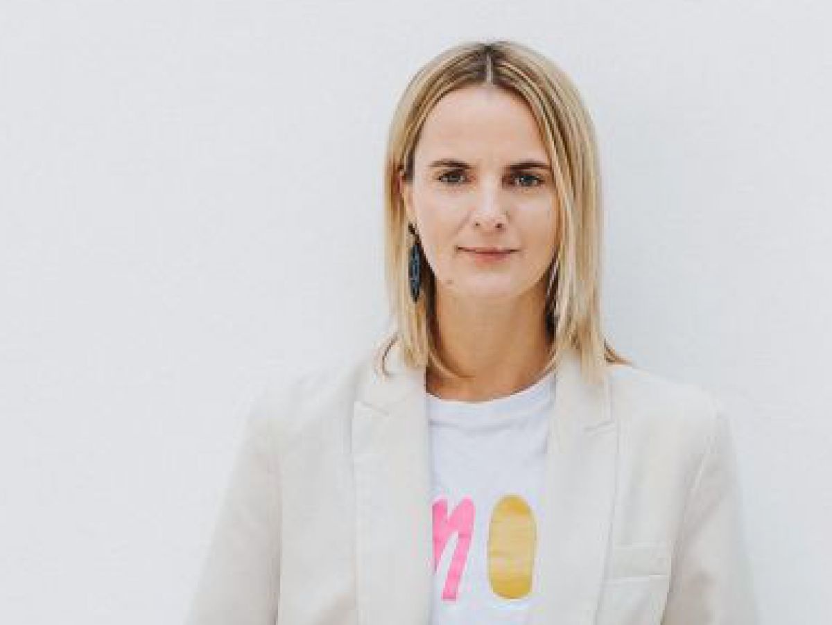 Schlanke Frau steht auf hellgrauem Betonboden und lehnt an weißer Wand. Ihr schulterlanges Haar ist blondiert, der Nachwuchs dunkel. Sie trägt eine weite weiße Hose, weiße Sneakers, ein weißes Shirt mit ein wenig neonfarbenem Aufdruck über der Brust und e