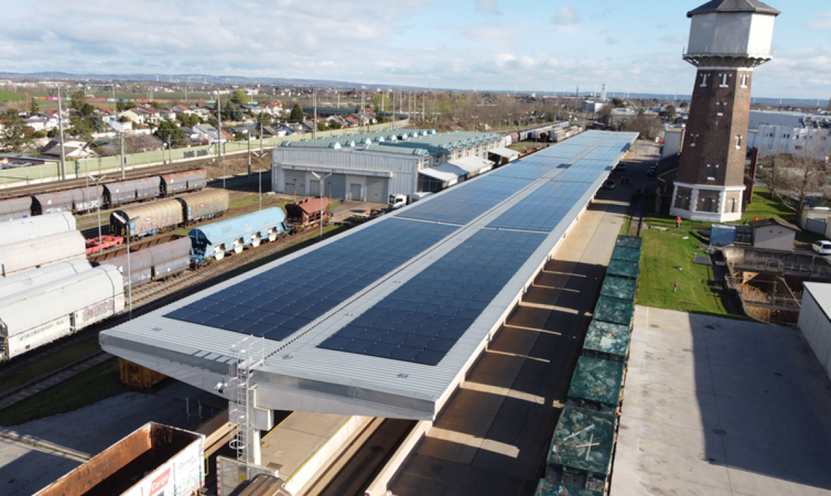 Blick von oben auf den Bahnhof in Süßenbrunn: In die Oberseite der Überdachung über den Bahnsteigen sind Photovoltaikzellen eingebaut. Auf den Abstellgleisen stehen Güterzüge, rechts im Bild ein historischer Speicherturm. Im Hintergrund die ebene bebaute 
