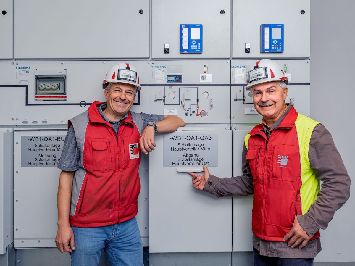 Zwei Herren in Arbeitsjacken und Schutzhelmen mit Firmenlogo von Baumit posieren vor Elektrokästen mit Stromzählern und Sicherungen.