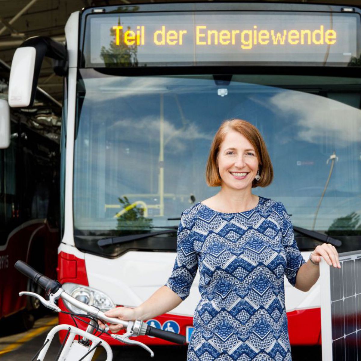 Gudrun Senk steht vor einem Bus und hält ein Fahrrad und ein PV-Modul in der Hand.