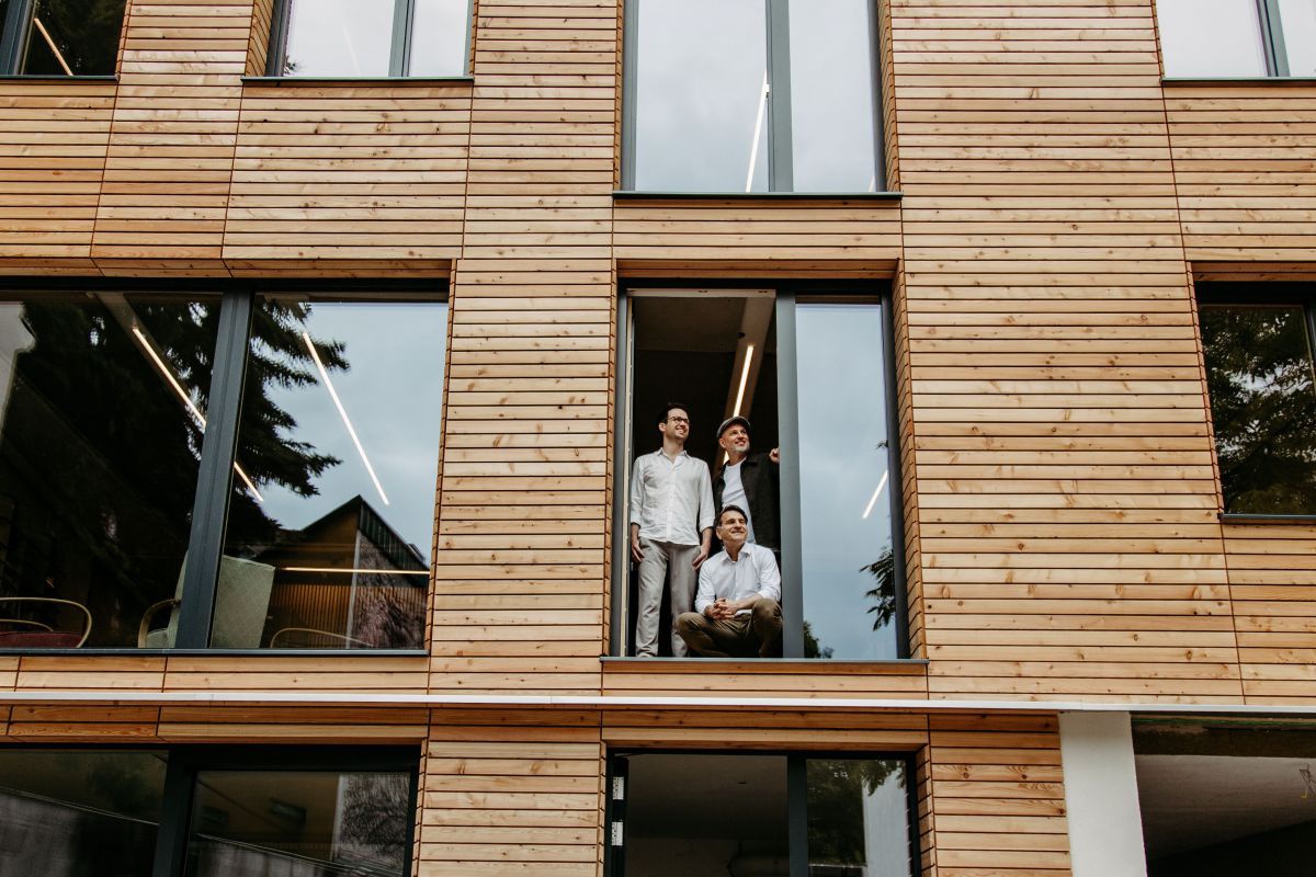 Drei Männer genießen den Ausblick aus dem Türrahmen einer offenen Balkontür eines Mehrparteienhauses, zwei stehen, einer hockt. Die Fassade ist mit waagerecht angebrachten, hellen Naturholzlatten verkleidet und hat viele Glasfenster in verschiedenen Größe