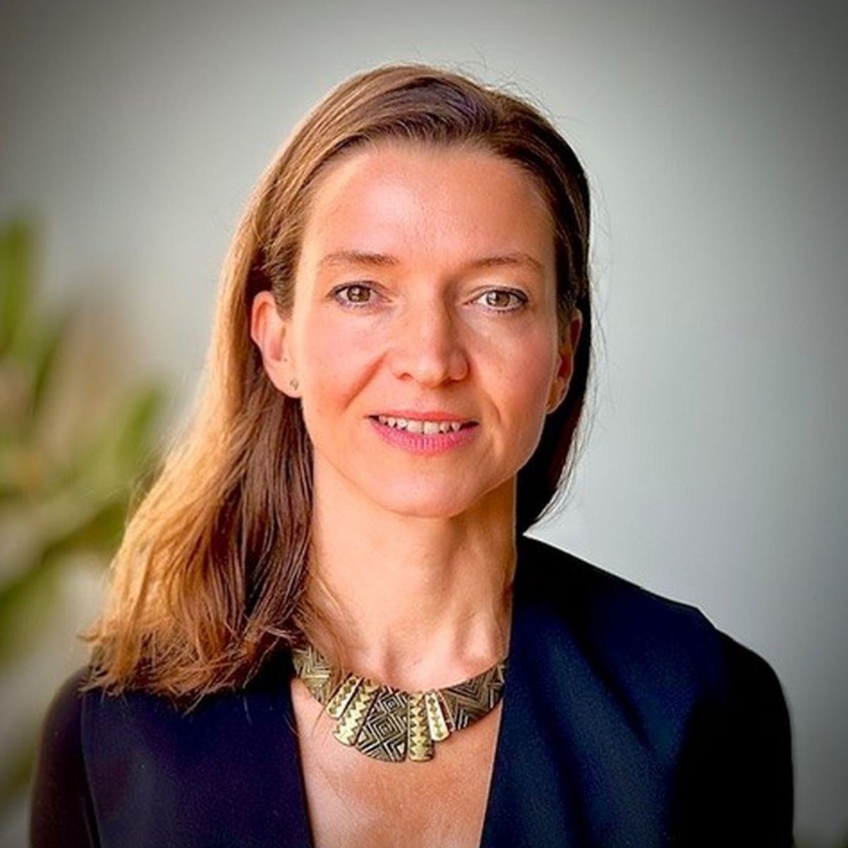 Portraitfoto einer Frau anfang vierzig mit langem, hellbraunem Haar, die in die Kamera lächelt. Sie trägt zum dunkelblauen Blazer einen Halsschmuck aus bronze- und messingfarbenen Metallplättchen mit eingravierten Ethno-Mustern.