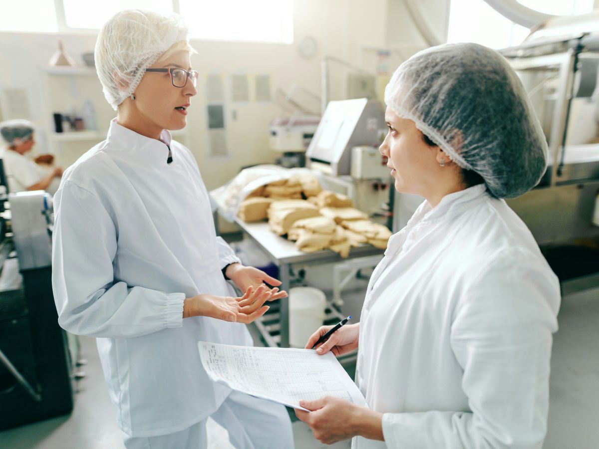 Zwei Mitarbeiter*innen in weißer Arbeitskleidung mit Haarnetzen stehen sich in einer Lebensmittelproduktionshalle im Gespräch gegenüber, eine hält ein Blatt Papier und einen Stift.