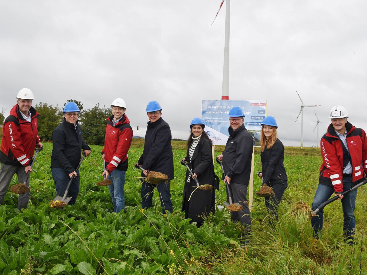 8 Personen stehen in einem Feld und halten Schaufeln mit Erde. Sie tragen weiße oder blaue Schutzhelme. Im Hintergrund sieht man Windräder.