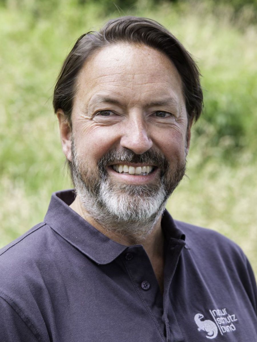 Ein Mann mittleren Alters mit gescheiteltem, braunem Haar und teilweise ergrautem Vollbart lächelt mit offenen Lippen ins Bild. Er trägt ein dunkelblau-graues T-Shirt mit dem Logo des Naturschutzbundes. Im Bildhintergrund sieht man verschwommen eine Wiese