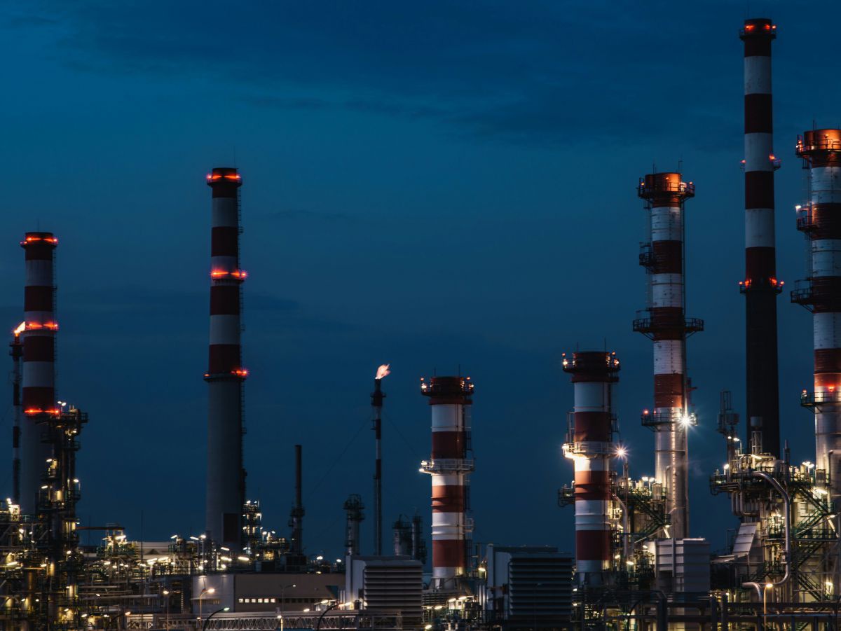 Blick auf ein Industriegebiet: Mehrere rot-weiß-gestreifte, mit kleinen Lichtern beleuchtete Schnornsteine in verschiedenen Höhen vor dunkelblauem Nachthimmel.