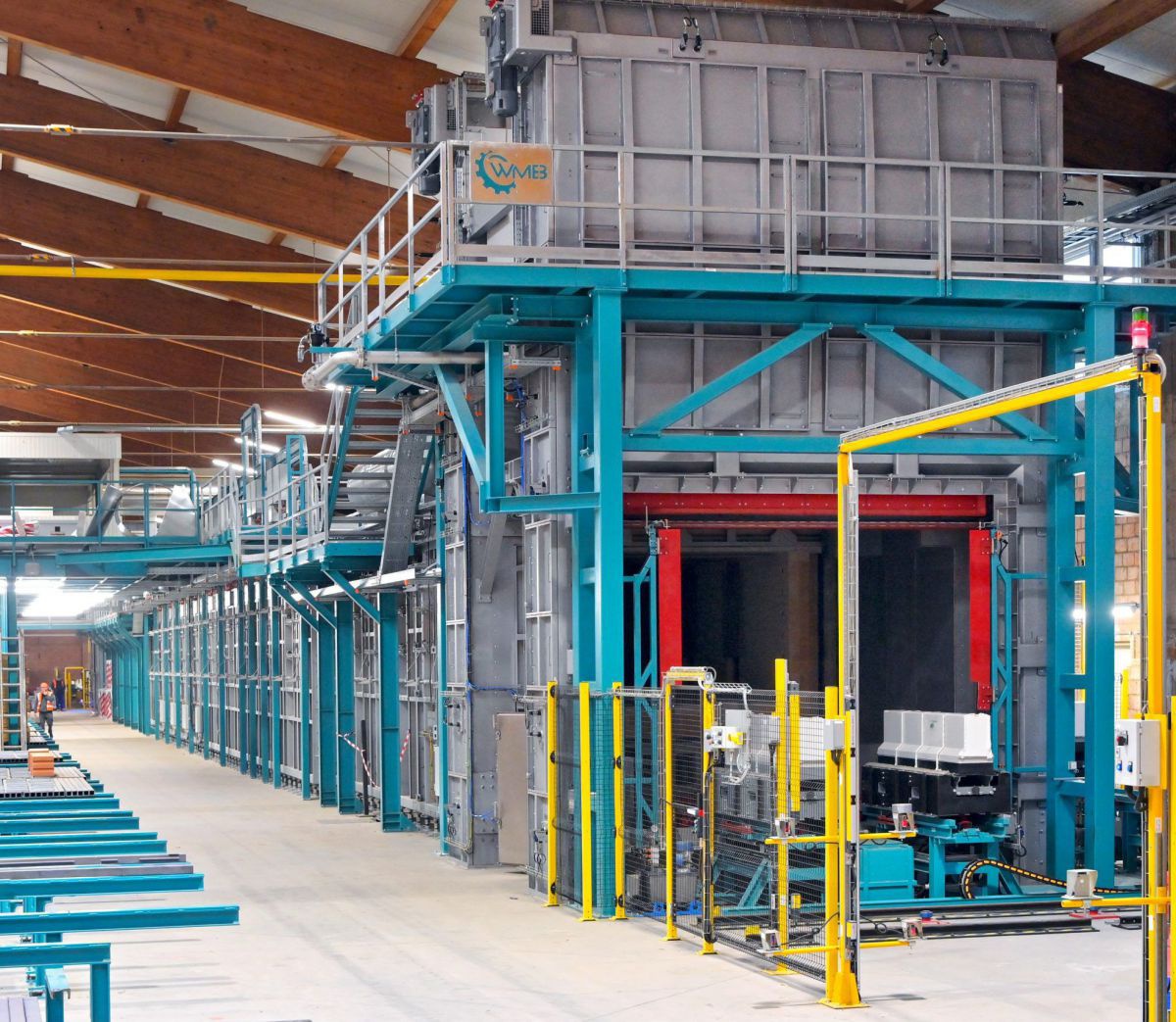 Auf dem Bild ist der riesige Elektroofen in der Werkshalle von Wienerberger zu sehen. Es sieht aus wie ein langgezogener, rechteckiger Container aus Beton, der von Metallgerüsten zusammengehalten wird. Die Gerüste sind in Grau, Türkis, Rot, Grün und Gelb 