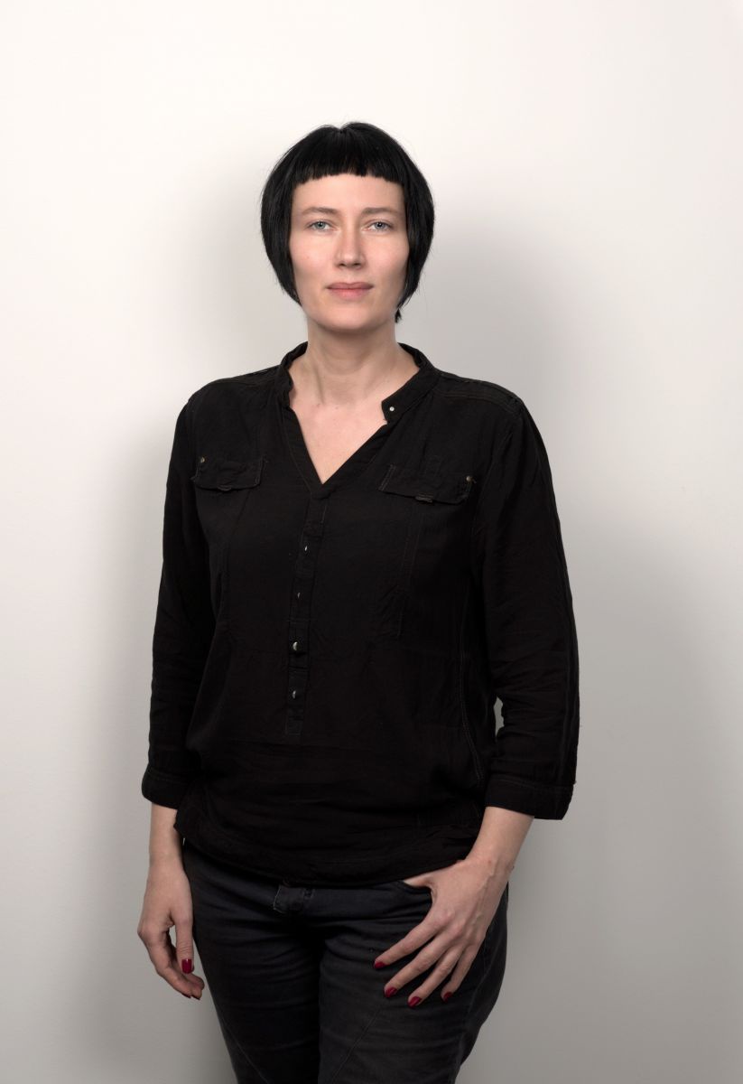 Martina Madner trägt ihr schwarzes Haar im kurzen Bob mit Micro Pony, dazu eine schwarze Hose und eine schware Hemdbluse.