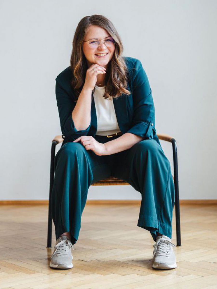 Eine junge Frau mit Brille und langem, gewellten Haar sitzt breitbeinig auf einem Stuhl in einem sonst leeren Raum mit Parkettboden und weißen Wänden. Sie trägt einen petrolfarbenen, weit geschnittenen Anzug und darunter ein weißes T-Shirt. Sie hat die El