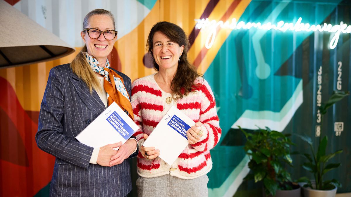 Zwei Frauen stehen vor der bunt bemalten Wand und präsentieren ihr Werk. Jede hält ein Exemplar des Buches in Händen.