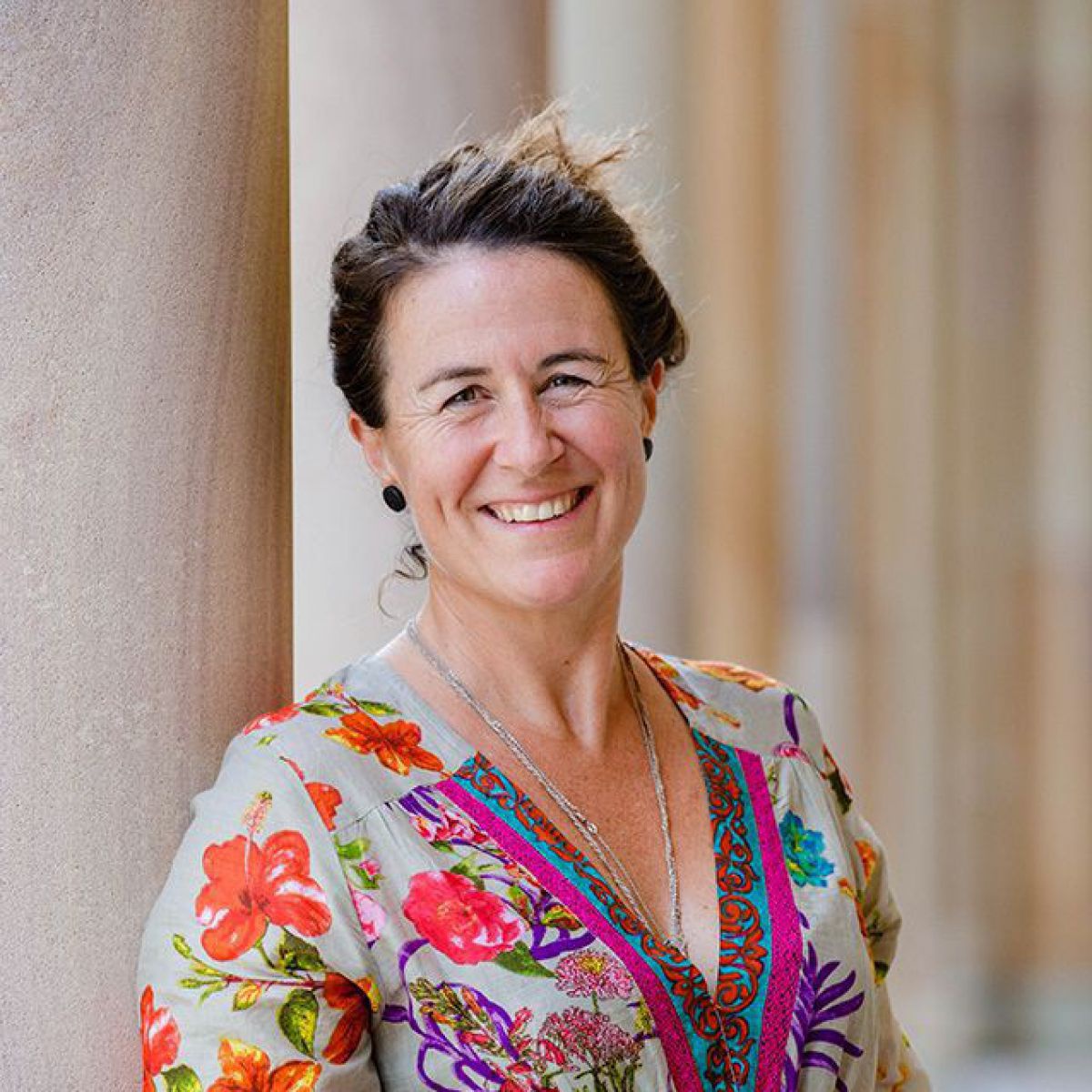 Eine Frau mittleren Alters im bunten Sommerkleid steht neben einer Säule und lächelt in die Kamera. Sie wirkt sehr erfrischend und glücklich.