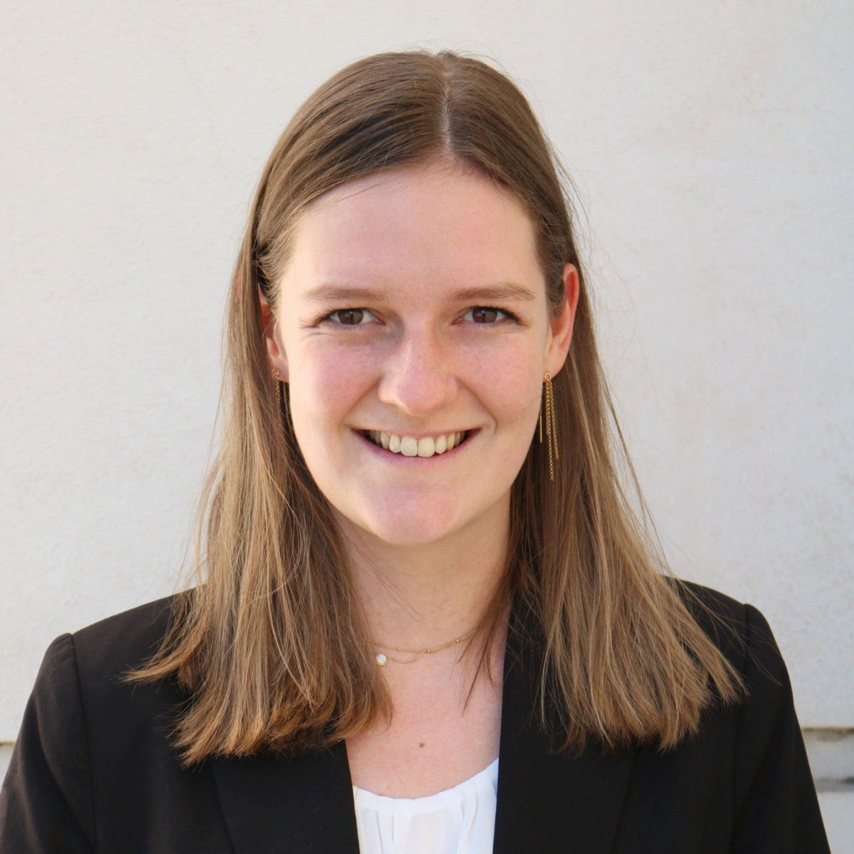 Portraitfoto von Rebecca Harbusch: Eine junge Frau mit dunkelblonden, schulterlangen Haaren lächelt in die Kamera. Sie trägt einen schwarzen Blazer über einer weißen Bluse mit Rundhalsausschnitt.