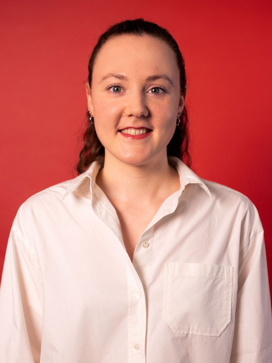 Portraitfoto von Leonie Marie Landmann: Eine junge Frau in weißem Herrenhemd und mit rot geschminkten Lippen steht vor einer roten Wand und lächelt in die Kamera. Ihr langes, gewelltes Haar trägt sie nach hinten gebunden.