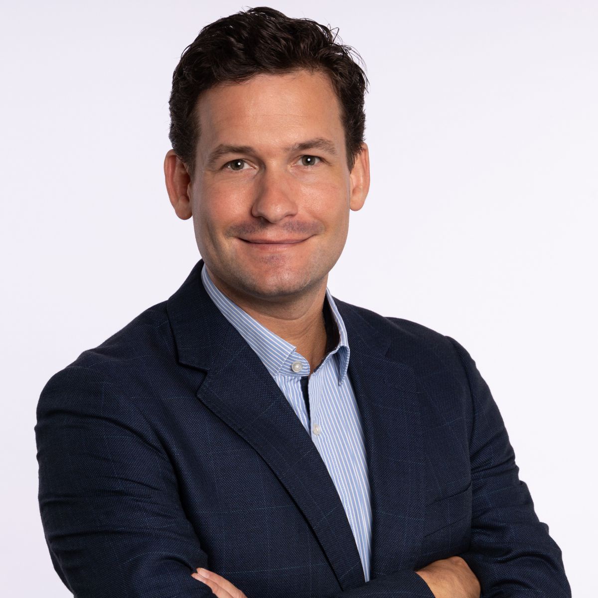 Portraitfoto von Mathias Nell: Ein junger Mann mit brauen kurzen Haaren und leichtem Bartschatten lächelt mit geschlossenen Lippen und hält die Arme vor der Brust verschränkt. Über einem hellblauen Hemd trägt er ein dunkelblaues Sakko.