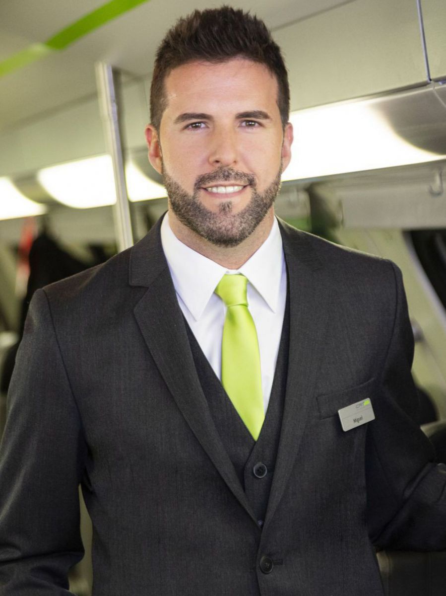 Miguel Centenero posiert in Dienstkleidung in einem Waggon des City Airport Train. Er trägt einen kurzen, dunklen Vollbart, sein dunkles Haar aufgestellt und kurz an den Seiten. Die Dienstkleidung von CAT ist ein anthrazitfarbener Anzug mit Weste darunter