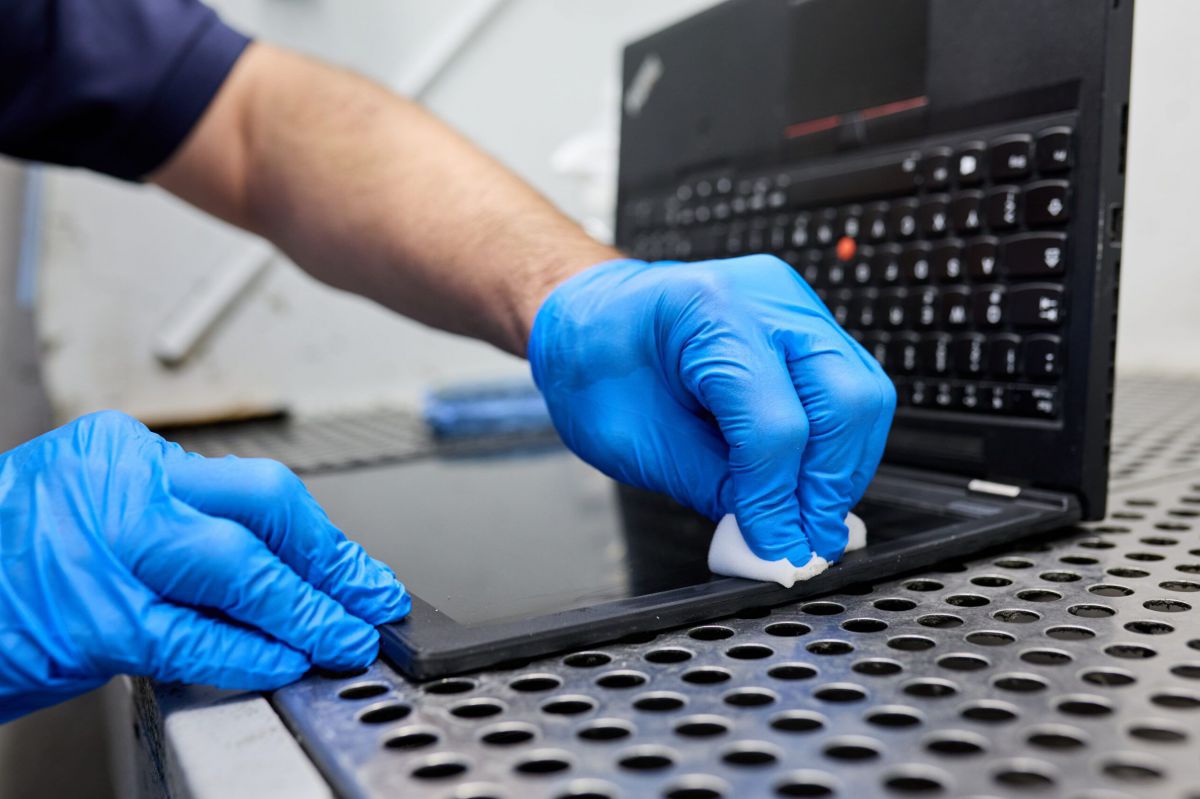 Ein Ausschnitt eines Laptops, der von einem Mitarbeiter, von dem man nur Unterarm bzw. Hände in blauen Gummihandschuhen sieht, mit einem Tuch poliert wird.