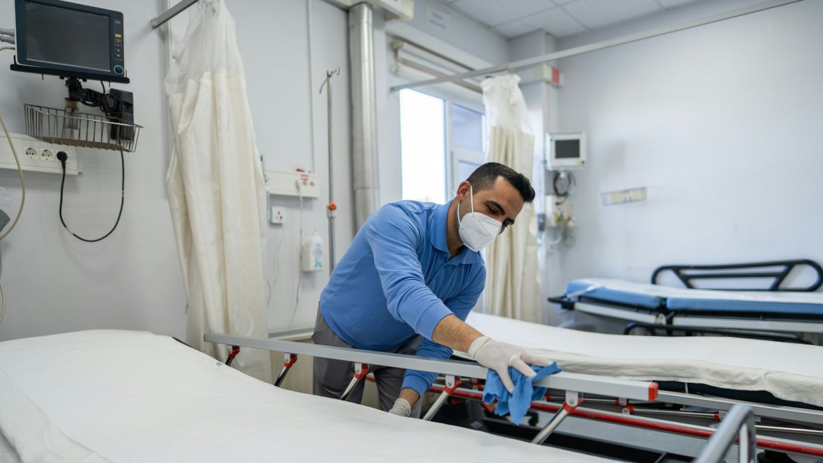 Ein Mann in hellblauer Arbeitskleidung trägt FFP2-Maske und Einweghandschuhe und wischt mit einem blauen Lappen das Gestell eines Krankenhausbettes ab.