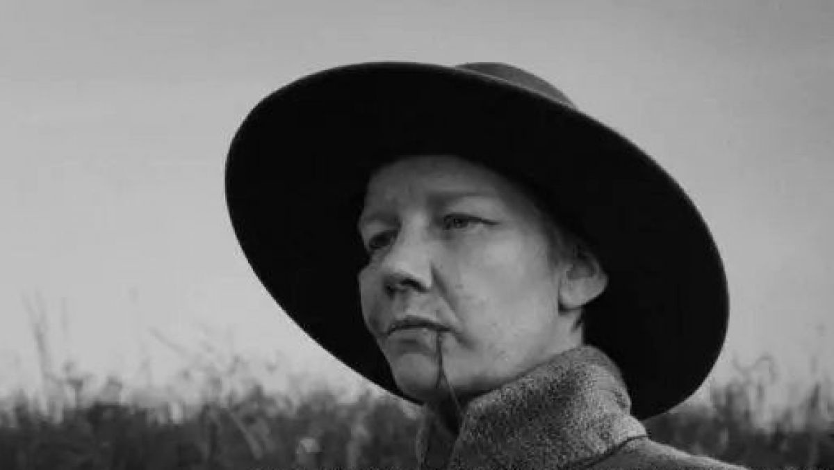 Bild in Schwarz-Weiß: Eine Frau mit Hut und hochgestelltem Mantelkragen in einer Landschaft mit hohem Gras und viel Himmel im Hintergrund. Zwischen den Lippen hat sie einen Halm oder Ähnliches.