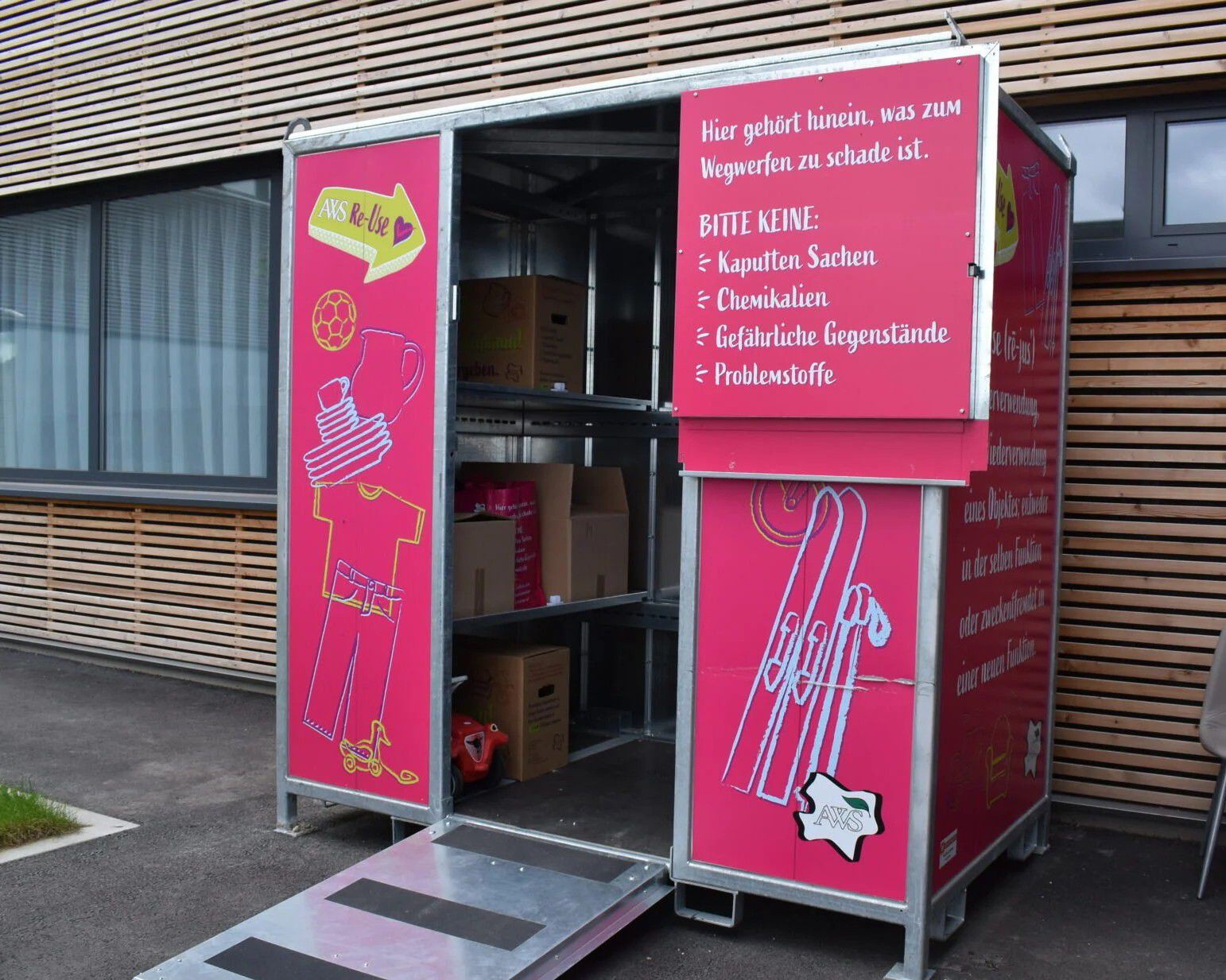 Ein kleiner, pinkfarbener Sammelcontainer für wiederverwendbare Gegenstände steht vor einem Gebäude mit Holzfassade. Die Tür des Containers ist geöffnet und gibt den Blick auf Regalböden mit Kartons im Inneren frei. Auf der Außenseite sind Illustrationen 