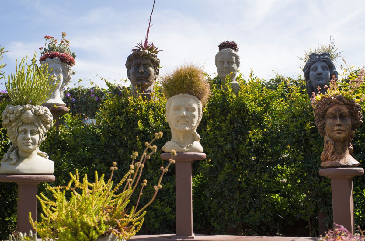 Steinerne Köpfe auf Podesten aus denen Pflanzen wachsen, in einem Park.