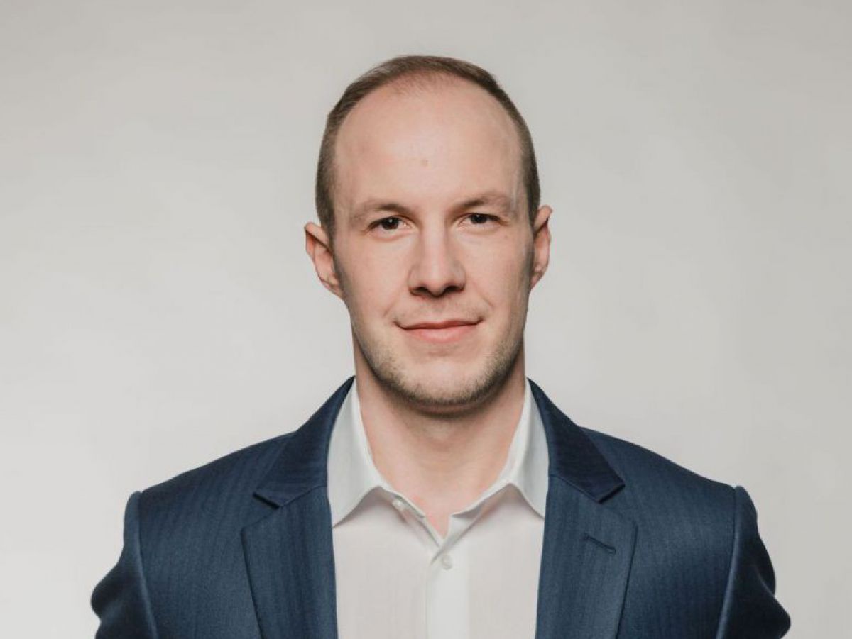 Portraitfoto eines großgewachsenen Mannes in den späten Dreißigern, er trägt einen blauen Anzug und ein weißes Hemd mit offenem Kragen ohne Krawatte.