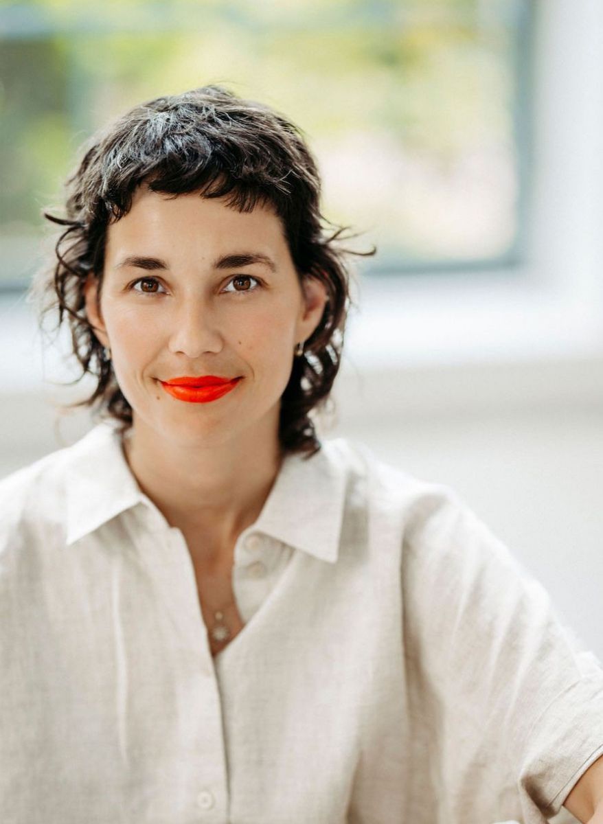 Portraitfoto einer jungen Frau mit dunklen, gewellten Haaren, dunklen Augen und korallenrot geschminkten Lippen. Sie trägt eine Hemdbluse aus naturweißem Leinen.