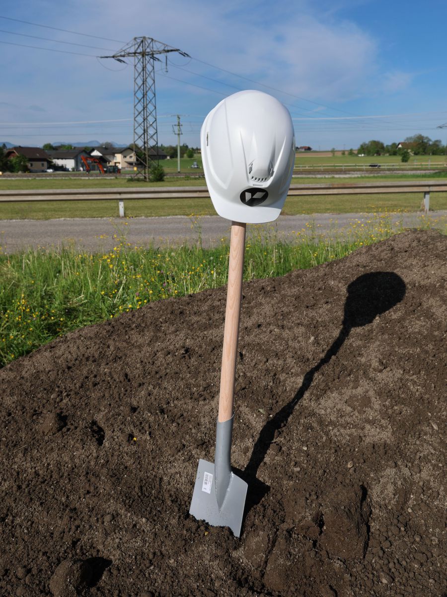 Ein Spaten steckt in einem Erdhaufen, an seinem Stiel hängt ein weißer Bauhelm. Im Hintergrund eine Landstraße, Stromleitungen, Landschaft und blauer Himmel.