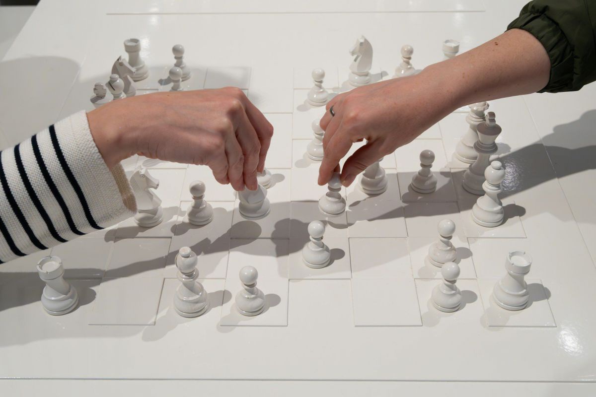 Zwei Personen spielen Schach, man sieht nur die Hände und Unterarme. Der Ärmel der Person links im Bild ist weiß-schwarz gestreift, jener der Person rechts im Bild ist schwarz. Das Schachspiel besteht ausschließlich aus weißen Figuren und Spielfeldern.
